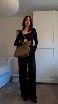 Μαύρη μπλούζα με μακριά μανίκια συνδυασμένη με μαύρο παντελόνι ψηλής μέσης· μοντέλο κρατά καφέ δερμάτινη τσάντα· ξύλινο πάτωμα και λευκή κλινοσκεπάσματα στο φόντο.