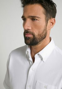 Camisa blanca de manga corta con cuello abotonado, que presenta un bolsillo delantero y una textura de tela sutil. Diseño limpio y sencillo.