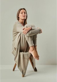 Beige Strick-Loungewear-Set mit langen Ärmeln und gerippten Bündchen, locker fallend, getragen auf einem Holzschemel sitzend. Glatte Textur, minimalistisches Design.