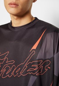 Svart sporttröja med hög krage, stort röd grafiskt tryck och ett slätt, texturerat tyg med prickiga mönster.