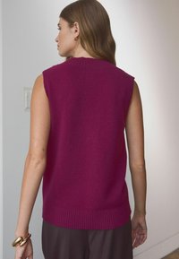 Fuchsia ärmelloser Pullover mit geripptem Saum und Ausschnitt, aus weichem Strickstoff, von hinten gezeigt, kombiniert mit braunen Hosen.