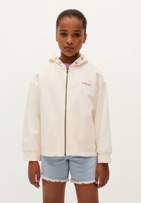 Crème-kleurige capuchon sweatshirt met een ritssluiting aan de voorkant, geribde manchetten en geborduurde branding. Gegdraagd met lichte denim shorts.