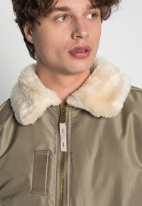 Chaqueta bomber en caqui con un suave cuello de pelaje sintético beige, cremallera dorada y detalles texturizados. Presenta un bolsillo parche en la parte frontal.