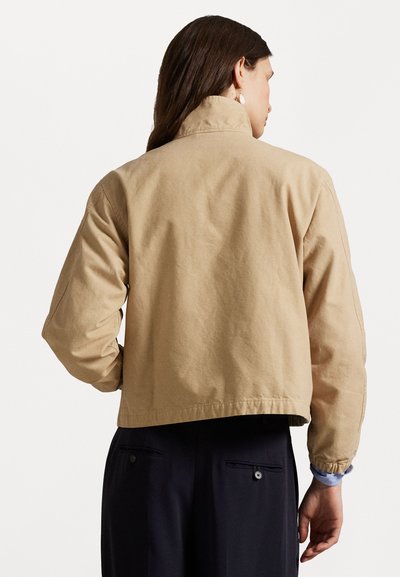 Beige verkürzte Jacke mit hohem Kragen und elastischen Bündchen, aus strukturiertem Baumwollstoff gefertigt und mit dezenten Stichdetails auf der Rückseite.