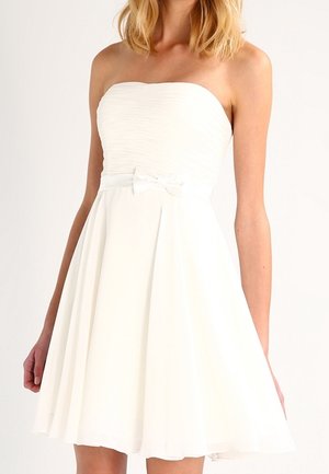 Olkaimeton valkoinen mekko, jossa on vartalonmyötäinen rypytetty yläosa, pieni rusetti vyötäröllä ja liehuva polvenpituinen hame, jota käyttää seisova henkilö.