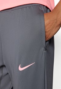 Grå treningsbukser laget av glatt, lett materiale. Har en rosa Nike-logo på venstre lår og en sidelomme.