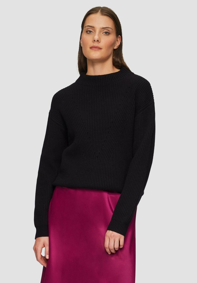Schwarzer, gerippter Strickpullover mit einem hohen Ausschnitt und einem V-förmigen Designdetail. Kombiniert mit einem glatten, glänzenden Magentorock.