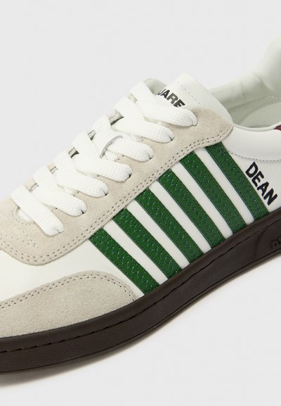 Baskets blanches avec des accents en suède gris, des rayures vertes et des lacets blancs. Présentent une semelle en caoutchouc noir et une étiquette "DEAN" sur le côté.