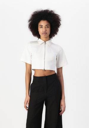 Korte, gecropte shirt met korte mouwen in roomkleur, voorzien van een rits aan de voorkant en een spitse kraag. Gecombineerd met hoge zwarte broeken.