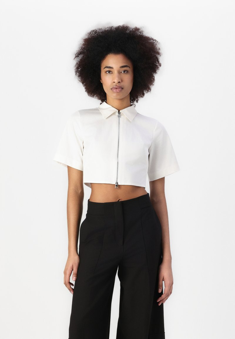 Camisa recortada de manga corta en color crema, con cremallera frontal y cuello puntiagudo. Combinada con pantalones negros de talle alto.