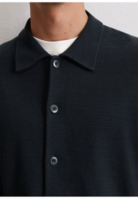 Dark navy shirt with a textured fabric, collar, and three black buttons. Close-up view focuses on neckline and button details.