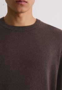 Nahaufnahme des unteren Gesichts und des Halses einer Person, die einen schlichten, dunkelbraunen Rundhals-Pullover vor einem schlichten weißen Wandhintergrund trägt.