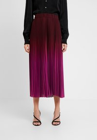 Plisseerock in Midi-Länge mit einem glatten, elastischen Bund, der einen Farbverlauf von tiefem Magenta zu hellem Pink zeigt. Kombiniert mit schwarzen Riemchensandalen.