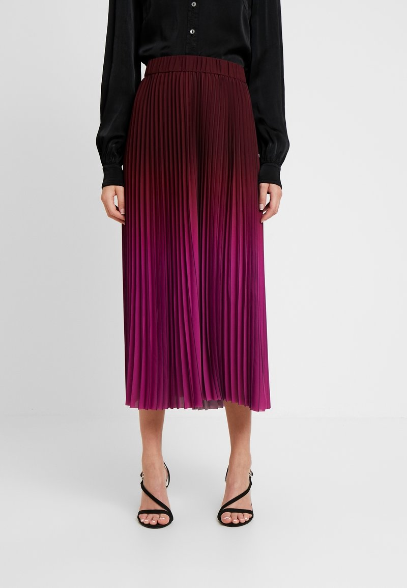 Plisseerock in Midi-Länge mit einem glatten, elastischen Bund, der einen Farbverlauf von tiefem Magenta zu hellem Pink zeigt. Kombiniert mit schwarzen Riemchensandalen.