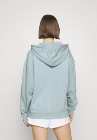 Světle modrá mikina s velkou kapucí, spadlými rameny a žebrovanými manžetami. Měkká tkanina, minimální detaily. Nosit přes bílé kraťasy.