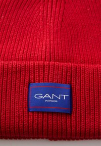 Red knitted beanie with ribbed texture and a blue rectangular label featuring "GANT SPORTSWEAR" in white text.