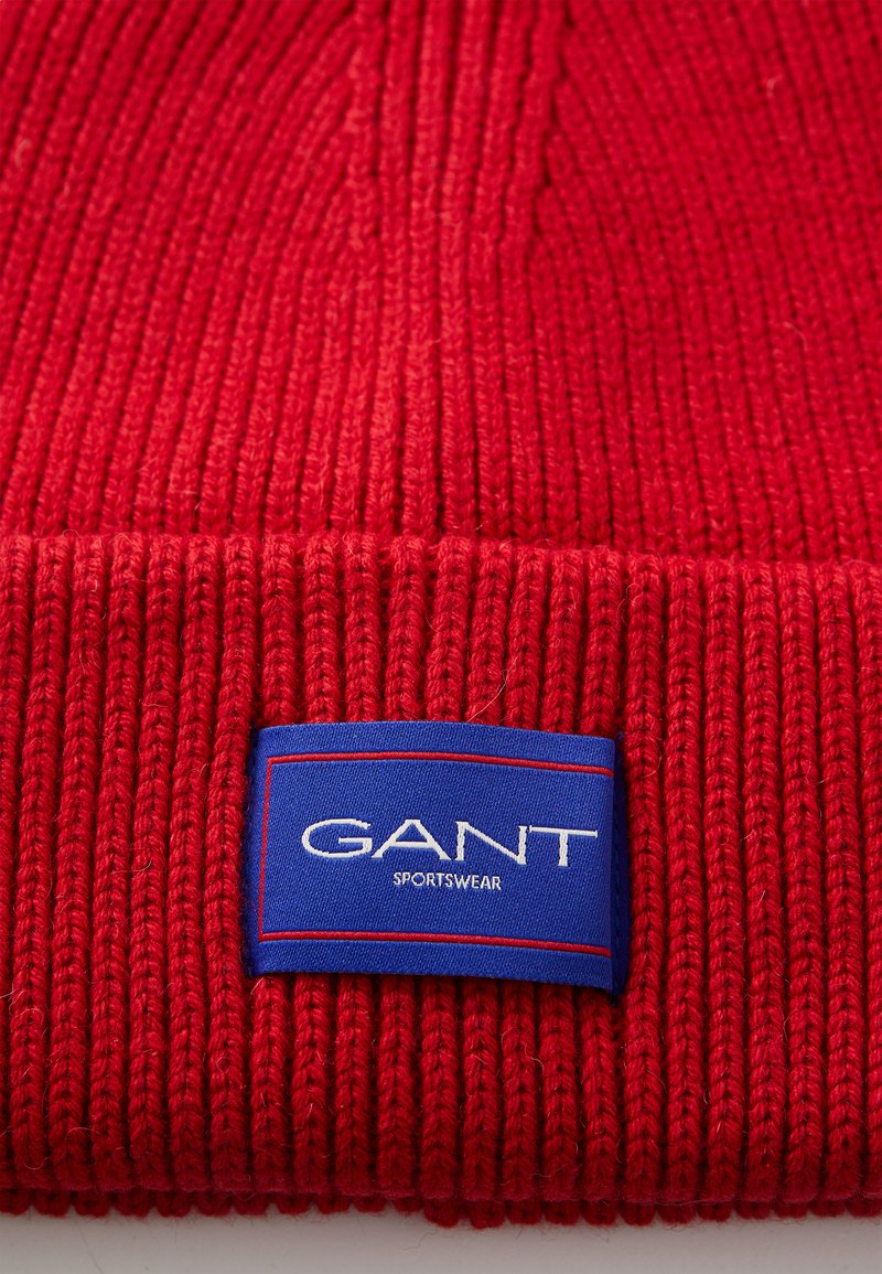 Rote gestrickte Mütze mit geripptem Muster und einem blauen rechteckigen Etikett, auf dem "GANT SPORTSWEAR" in weißem Text steht.