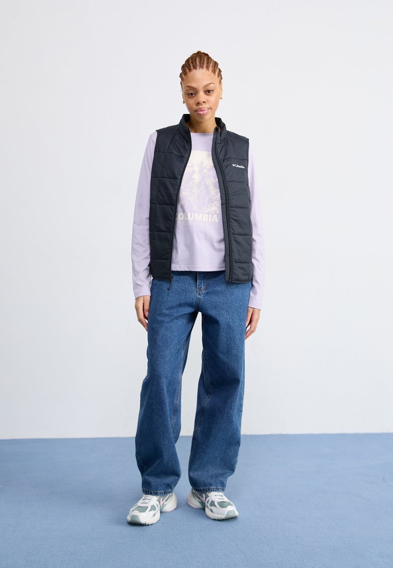 Chaleco negro acolchado sobre una camisa de manga larga de color púrpura claro con un estampado gráfico, combinado con jeans azules anchos y zapatillas grises.