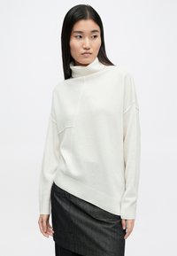 Κρεμ χρώματος πουλόβερ με ψηλό λαιμό, φαρδιά εφαρμογή, ασύμμετρο τελείωμα, ραβδωτά μανίκια και μέση. Μαλακή υφή και μινιμαλιστικός σχεδιασμός.