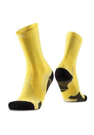Een paar gele sportsokken met zwarte hiel- en teenstukken, ribbelstructuur en wit logo vlakbij de bovenste boord, getoond op voetvormige mallen.