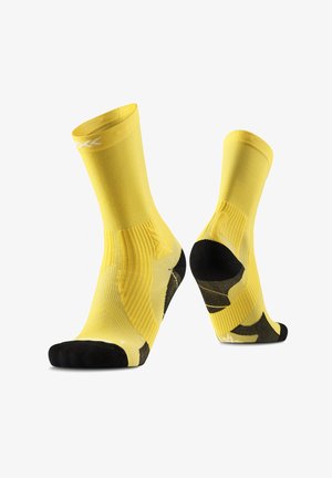 Paire de chaussettes de sport jaunes avec talon et pointe noirs, texture côtelée et logo blanc près du bord supérieur, présentées sur des formes en forme de pied.