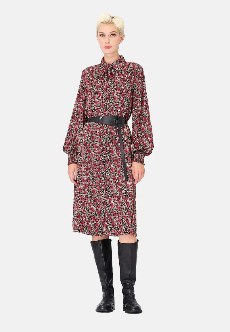 Robe florale rouge avec un col et des manches longues, cintrée à la taille par une ceinture noire. Modèle portant des bottes noires jusqu'aux genoux.