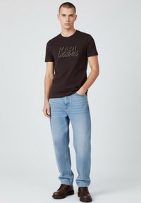 Czarna bawełniana koszulka z złotym logo "KARL LAGERFELD", zestawiona z jasnoniebieskimi dżinsami i brązowymi sznurowanymi butami. Prosty, codzienny strój.