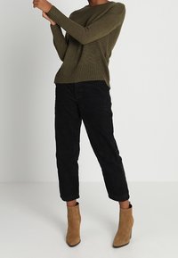 Pull tricot vert olive avec une finition texturée, associé à un pantalon court en velours côtelé noir et des bottines marron clair.