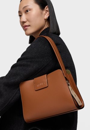 Femme en manteau noir portant un sac à bandoulière TOUS en cuir marron avec des ferrures dorées et une étiquette en cuir beige.