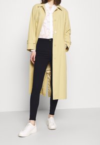 Trench coat amarillo claro sobre una camisa blanca con estampado, combinado con leggings negros y zapatillas blancas. Texturas suaves y corte relajado.