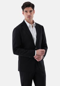 Schwarze taillierte Anzugjacke mit Ein-Knopf-Verschluss, schlanker Passform und Reverskragen. Trägt man über einem weiß-blau gestreiften Hemd.