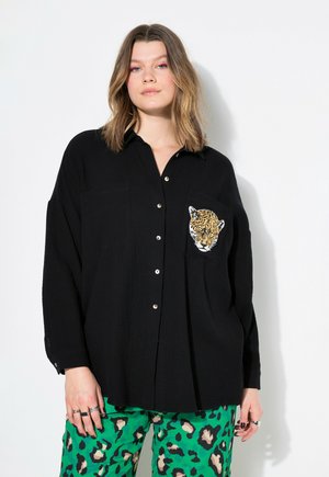Maglietta nera a maniche lunghe con chiusura a bottoni e tasca, caratterizzata da un ricamo a testa di leopardo. Tessuto strutturato. Modello abbinato a pantaloni verdi con stampa leopardata.
