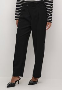 Pantalons noirs ajustés avec une taille haute, fermeture à double bouton, plis à l'avant et jambes droites, associés à des mules noires scintillantes.