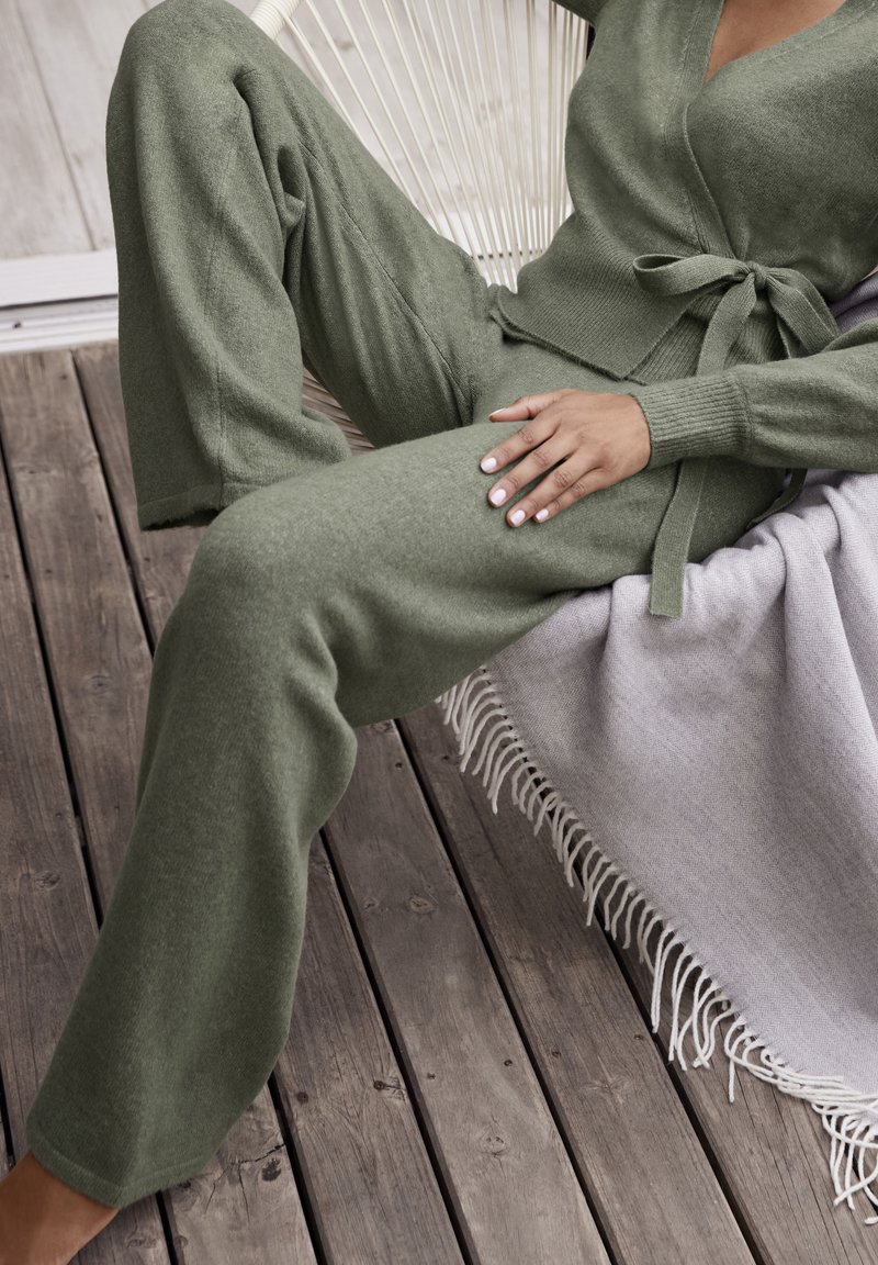 Olivgrünes gestricktes Loungewear-Set mit einem Wickeldesign, einer gebundenen Taille, weiten Hosenbeinen und gerippten Bündchen, vor einem hellgrauen Handtuch.