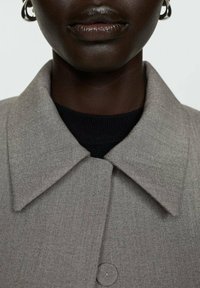 Cappotto grigio intrecciato con un ampio colletto, caratterizzato da una finitura testurizzata e un dettaglio con bottone sul davanti, indossato sopra una maglietta nera.