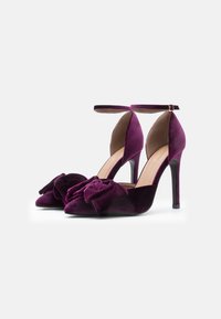 Salto alto de veludo roxo com bico fino, apresentando um grande detalhe de laço na parte da frente e uma tira delicada no tornozelo.