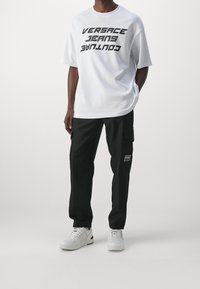 Bílý tričko s výrazným černým textem "VERSACE JEANS COUTURE", v kombinaci s černými cargo kalhotami se bočními kapsami a logem, doplněné o bílé tenisky.