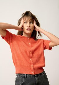 Kortärmad cardigan i livlig orange stickning, med rund hals och knappar fram. Mjuk textur och avslappnad passform.