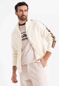 Veste à zip crème avec des rayures marron sur les manches, portée sur un t-shirt beige comportant un texte noir. Le pantalon est en tissu léger, assorti à la veste.