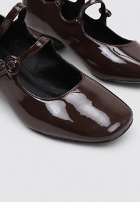 Zapatos Mary Jane de charol marrón con punteras cuadradas, una correa redondeada con hebilla y detalles recortados en los laterales.