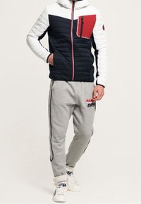 Polstret jakke i marineblå og hvid med røde detaljer, lynlåslomme, parret med grå sweatpants med sidelister og logo, båret med hvide sneakers.