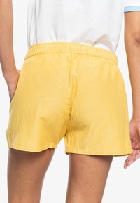 Personne portant un short jaune à taille élastique et une chemise blanche, avec une main dans la poche du short, vue de dos.