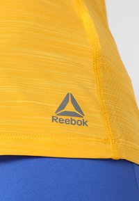 Gul idrottströja med fukttransporterande textur, korta ärmar, med en grå Reebok-logotyp på vänster nedre sida.