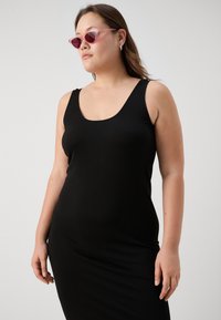 Svart ribbestrikket tankkjole med scoop-neck og brede stropper. Modellen har på seg rosa katteeøyne-solbriller og har langt hår.