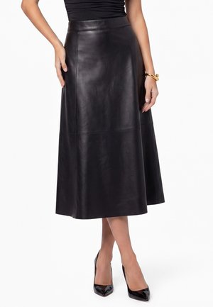 Zwarte leren midi-rok met hoge taille, gedragen met zwarte pumps met puntige neus en een gouden armband om de rechterpols.