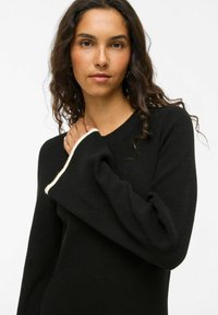 Černý svetr s výstřihem do V a dlouhými rukávy, zdobený kontrastním krémovým lemováním na manžetách. Materiál působí měkce, s texturovaným pleteným povrchem.