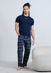 Άνδρας με ράστα στα μαλλιά στέκεται ξυπόλυτος φορώντας μπλε ναυτικό μπλουζάκι και μπλε καρό πιτζάμες δίπλα σε μια λευκή υφή πολυθρόνα.