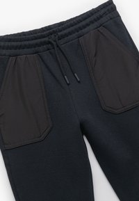 Pantalones de chándal en azul marino oscuro con cintura elástica, cordón ajustable y dos grandes bolsillos angulados en tejido oscuro contrastante. Textura suave, corte relajado.