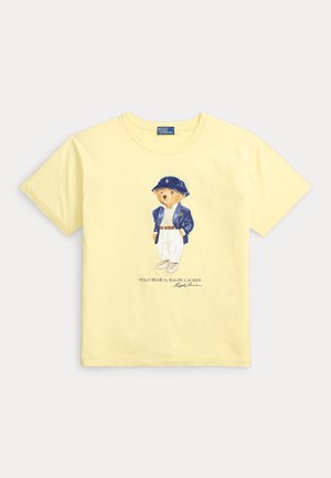 T-shirt jaune à manches courtes avec un ours portant un chapeau bleu, une veste bleue, un pantalon blanc et des baskets, avec le texte Polo Bear by Ralph Lauren en dessous.