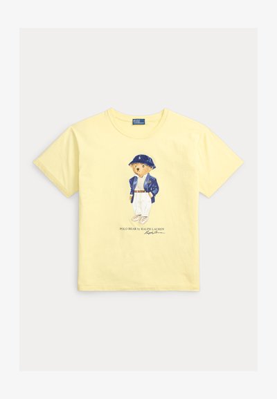 Κίτρινο T-shirt με κοντά μανίκια, που απεικονίζει μια αρκούδα με μπλε καπέλο, μπλε μπουφάν, λευκό παντελόνι και αθλητικά παπούτσια, με κείμενο Polo Bear by Ralph Lauren από κάτω.